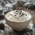 A bowl of creamy snow cream topped with chocolate chips, crushed cookies, and syrup, sitting on a rustic wooden table surrounded by soft snowflakes and snow-covered trees in a wintery landscape.